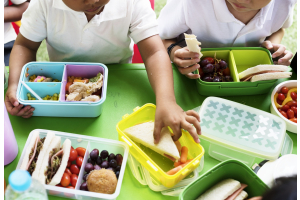 Children in white shirts enjoy lunch with colourful lunchboxes on a green table. Boxes contain sandwiches, fruits, and vegetables, evoking a playful, cheerful tone.